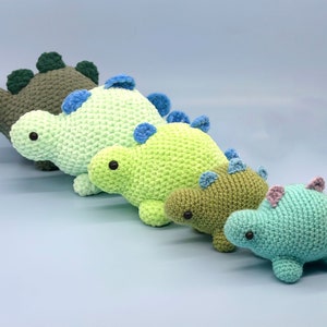 May include: Four crocheted dinosaur toys in different sizes and colours. The dinosaurs are green, light green, and blue. They have black eyes and are arranged in a line from largest to smallest.