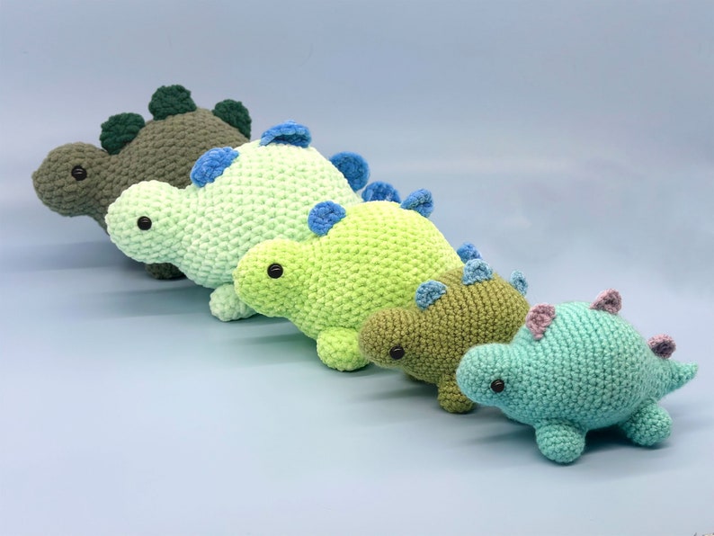 May include: Four crocheted dinosaur toys in different sizes and colours. The dinosaurs are green, light green, and blue. They have black eyes and are arranged in a line from largest to smallest.