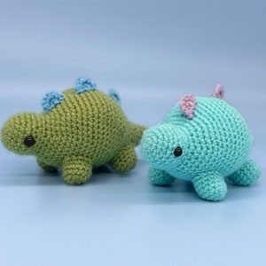 May include: Two crocheted dinosaur toys. One is green with blue accents and the other is blue with pink accents.