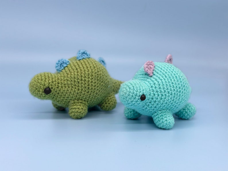 May include: Two crocheted dinosaur toys. One is green with blue accents and the other is blue with pink accents.
