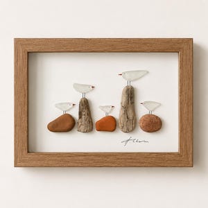 May include: A framed wall art piece featuring five stylised birds created from white sea glass and small metal wire legs. The birds are perched on various brown and orange river stones and pieces of driftwood. A signature is visible in the corner.