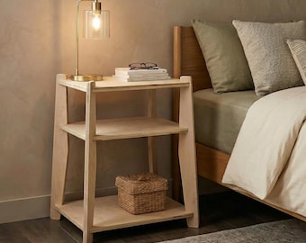 May include: A light-colored wooden nightstand with three shelves. A gold-toned lamp with a glass shade sits on top, next to a stack of books and eyeglasses. A woven basket is on the bottom shelf. The nightstand is next to a bed with neutral-colored bedding.