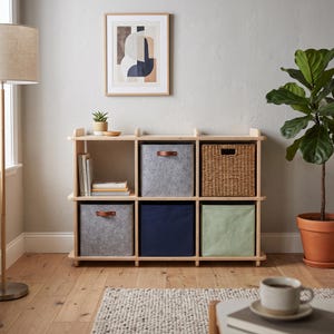 May include: A light-colored wooden storage unit with six cubby compartments. The unit contains two grey felt bins with brown leather handles, one woven wicker basket, one dark blue fabric bin, one light green fabric bin, and a stack of books.