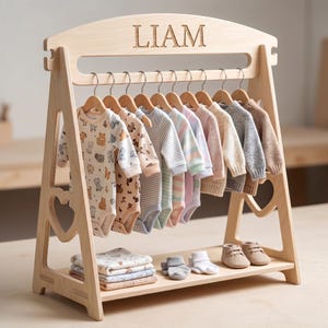 Könnte beinhalten: Ein heller Holzkleiderständer mit dem eingravierten Namen LIAM an der Oberseite. Der Ständer hält mehrere Babybodys auf Kleiderbügeln, während gefaltete Stoffe und ein Paar kleine beige Schuhe auf der unteren Ablage des Gestells liegen.