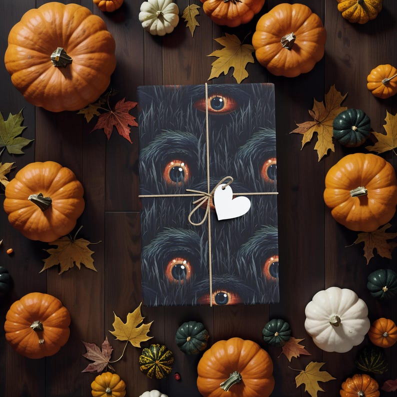May include: A black and white wrapping paper with a spooky eye pattern. The paper is tied with twine and a white heart-shaped tag. The paper is surrounded by orange pumpkins and other fall decorations.