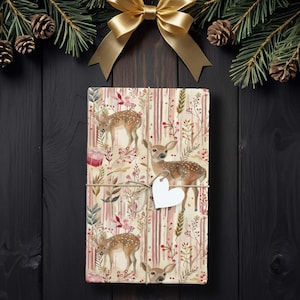 May include: A wrapped gift with deer and floral patterned paper, tied with twine and a white heart-shaped tag. The gift is decorated with a gold bow and pine branches with pinecones, set against a dark wood background.