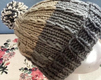 Può includere: Un berretto lavorato a mano a maglia nei toni del grigio, beige e crema. Il cappello presenta un design a coste e un soffice pompon sulla parte superiore. Il cappello è esposto su una testa di manichino bianca.