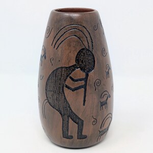 May include: A brown vase with a tapered cylindrical shape featuring a dark etched design of a flute player and several small horned animals. The surface is decorated with black engraved figures and small spiral patterns across the exterior.