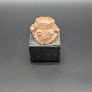 May include: A small, terracotta-colored sculpture of a face with closed eyes and a hat. The face has a serene expression and is mounted on a small, black, rectangular block. The sculpture is likely an ancient artifact or a replica.