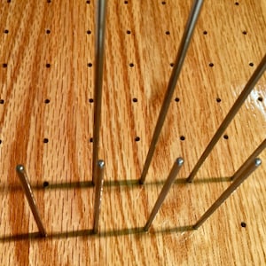 May include: A wooden board with a grid of holes, each with a metal pin inserted. The pins are arranged in a pattern, creating a decorative and functional design.