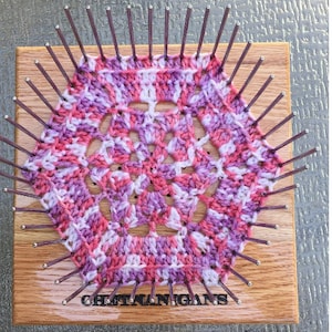 May include: A pink and white crocheted doily is being made on a wooden loom with metal pegs. The loom has the text "Gretna Nigans" printed on it.
