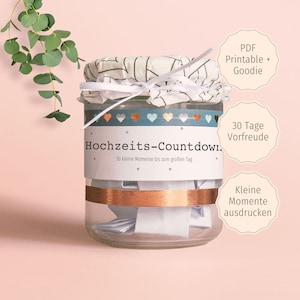 Puede incluir: Un tarro de vidrio con tapa blanca y una etiqueta azul y blanca que dice "Hochzeits-Countdown" con el texto "30 kleine Momente bis zum großen Tag". El tarro está lleno de pequeños trozos de papel doblados. Hay tres etiquetas en el tarro: "PDF Printable + Goodie", "30 Tage Vorfreude" y "Kleine Momente ausdrucken".