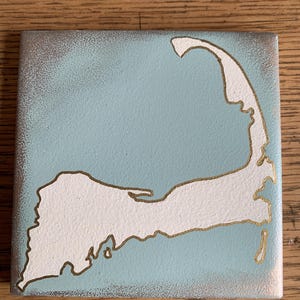 May include: A light blue square tile with a white outline of Cape Cod painted on it. The outline is accented with a thin gold line.