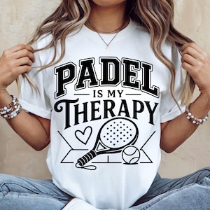 May include: A white short-sleeve t-shirt featuring black graphic text that reads PADEL IS MY THERAPY. The design includes a stylized illustration of a padel racket, a ball, and a heart, all centered on the front of the garment.