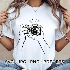 Può includere: T-shirt bianca con un'illustrazione nera di una fotocamera tenuta in mano. Il testo "SVG - JPG - PNG - PDF - EPS" è in basso. La maglietta è a girocollo.