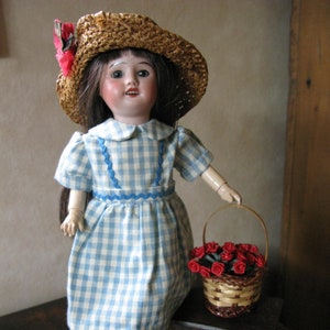Peut inclure: Une poupée en porcelaine portant une robe à carreaux bleus et blancs et un chapeau de paille avec un ruban rouge. La poupée tient un panier de roses rouges.