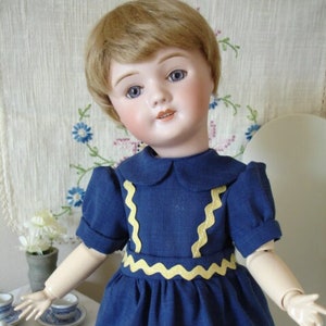 May include: A porcelain doll with blonde hair wearing a blue dress with yellow trim. The doll has blue eyes and a painted smile.