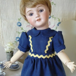 May include: A porcelain doll with blonde hair wearing a blue dress with yellow trim. The doll has blue eyes and a painted smile.