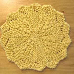 Puede incluir: Un artículo tejido a mano, redondo, amarillo, con un borde festoneado. La pieza tejida tiene un patrón de rayos de sol que irradia desde el centro. El artículo es probablemente un accesorio de cocina, como un salvamanteles o un paño de cocina.