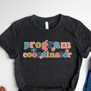 May include: A dark heather grey t-shirt with the text "program coordinator" in colorful letters with flower accents.