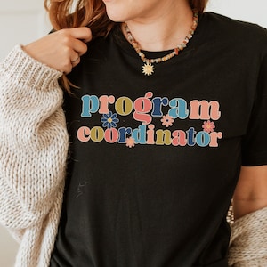 May include: Black t-shirt with the text "program coordinator" in colorful retro letters with flowers. The shirt is worn by a person with a cardigan and jeans.