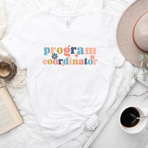 May include: White t-shirt with the text "program coordinator" in colorful retro lettering with flowers.