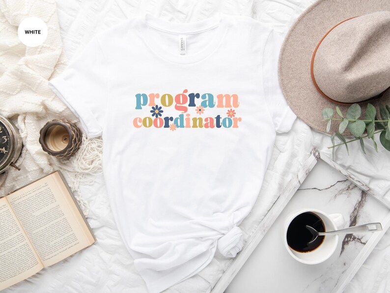 May include: White t-shirt with the text "program coordinator" in colorful retro lettering with flowers.