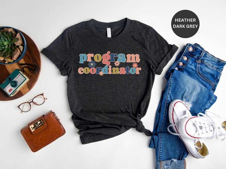 May include: A dark heather grey t-shirt with the text "program coordinator" in colorful letters with flower accents.
