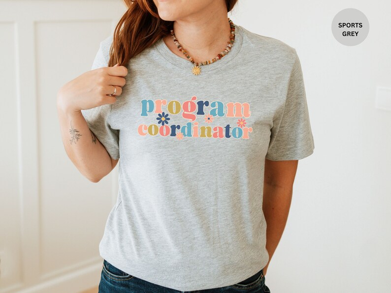 May include: A grey t-shirt with the text "program coordinator" in colorful letters with flowers. The shirt is a sports grey color.