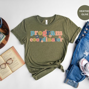 May include: A green t-shirt with the text "program coordinator" in colorful letters with floral accents. The shirt is on a white background with a pair of blue jeans and white sneakers.