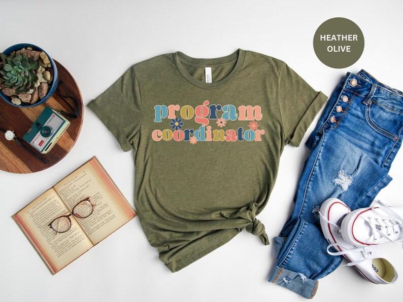 May include: A green t-shirt with the text "program coordinator" in colorful letters with floral accents. The shirt is on a white background with a pair of blue jeans and white sneakers.