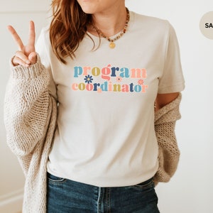 May include: A white t-shirt with the text "program coordinator" in colorful letters with flowers. The text is in a retro style font.