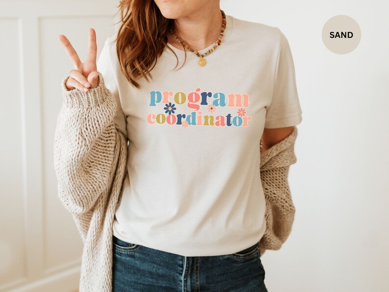 May include: A white t-shirt with the text "program coordinator" in colorful letters with flowers. The text is in a retro style font.