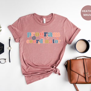 May include: A heather mauve t-shirt with the text "program coordinator" in colorful retro lettering with floral accents.