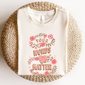 May include: A cream-colored t-shirt with a floral design and the text "Your Words Matter" in brown lettering with a pink heart.
