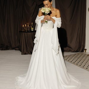 May include: A white wedding dress with an off-the-shoulder neckline and flowing skirt. The bride holds a bouquet of white flowers. The dress features a fitted bodice and long, draped sleeves. The background includes brown curtains and a small table.