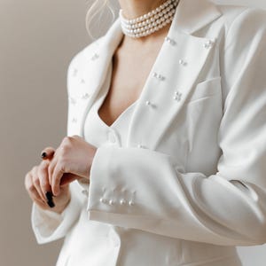 Puede incluir: Una chaqueta blanca con ribete de perlas y un collar de perlas. La chaqueta tiene cierre de doble botonadura y una silueta ajustada.
