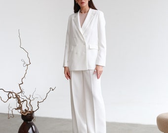 May include: A woman wearing a white double-breasted blazer and matching wide-leg pants. She is wearing white sunglasses and has her hair pulled back.