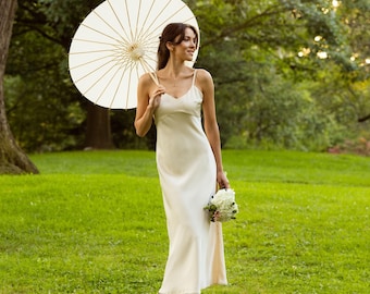 May include: A woman in a white dress holds a white bouquet of flowers and a white parasol in a green grassy field.