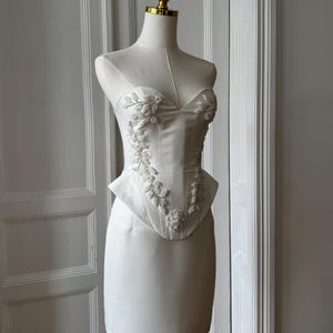 May include: A white strapless dress with floral embroidery and beading. The bodice features a sweetheart neckline and a peplum waist. The dress is displayed on a mannequin with a gold-colored head. The dress is made of a smooth, satin-like fabric.