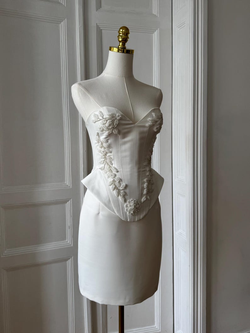 May include: A white strapless dress with floral embroidery and beading. The bodice features a sweetheart neckline and a peplum waist. The dress is displayed on a mannequin with a gold-colored head. The dress is made of a smooth, satin-like fabric.