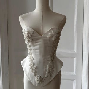 May include: Ivory-colored corset with a sweetheart neckline, displayed on a mannequin. The bodice is embellished with delicate beaded floral and leaf patterns. The corset's design features a structured silhouette, showcasing a classic bridal or formal wear style.
