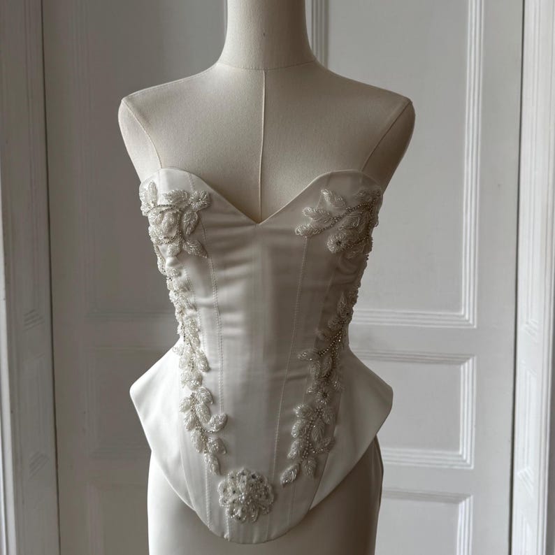 May include: Ivory-colored corset with a sweetheart neckline, displayed on a mannequin. The bodice is embellished with delicate beaded floral and leaf patterns. The corset's design features a structured silhouette, showcasing a classic bridal or formal wear style.