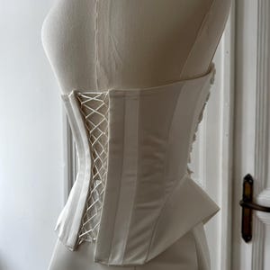 May include: Ivory-colored corset with a lace-up back, displayed on a mannequin. The corset features vertical boning and a structured design. The fabric has a satin sheen, and the garment is likely for formal wear or bridal attire.