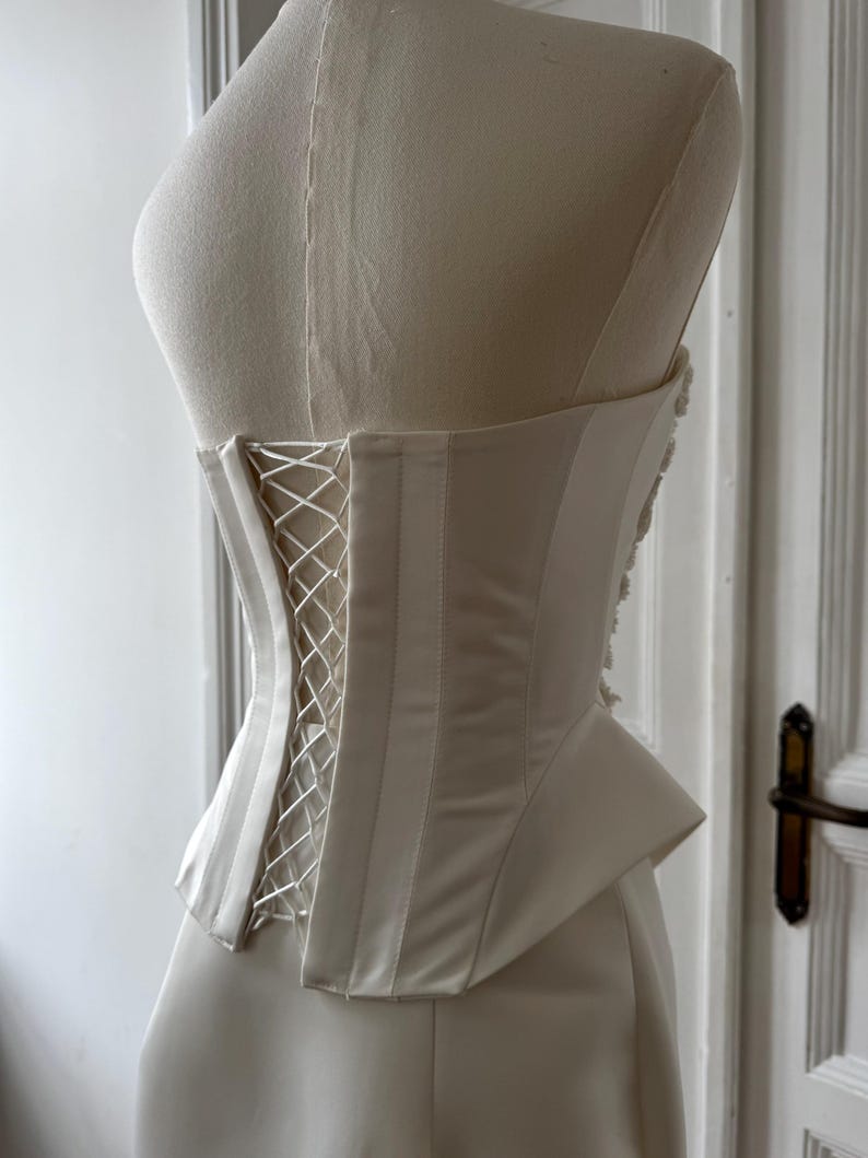 May include: Ivory-colored corset with a lace-up back, displayed on a mannequin. The corset features vertical boning and a structured design. The fabric has a satin sheen, and the garment is likely for formal wear or bridal attire.