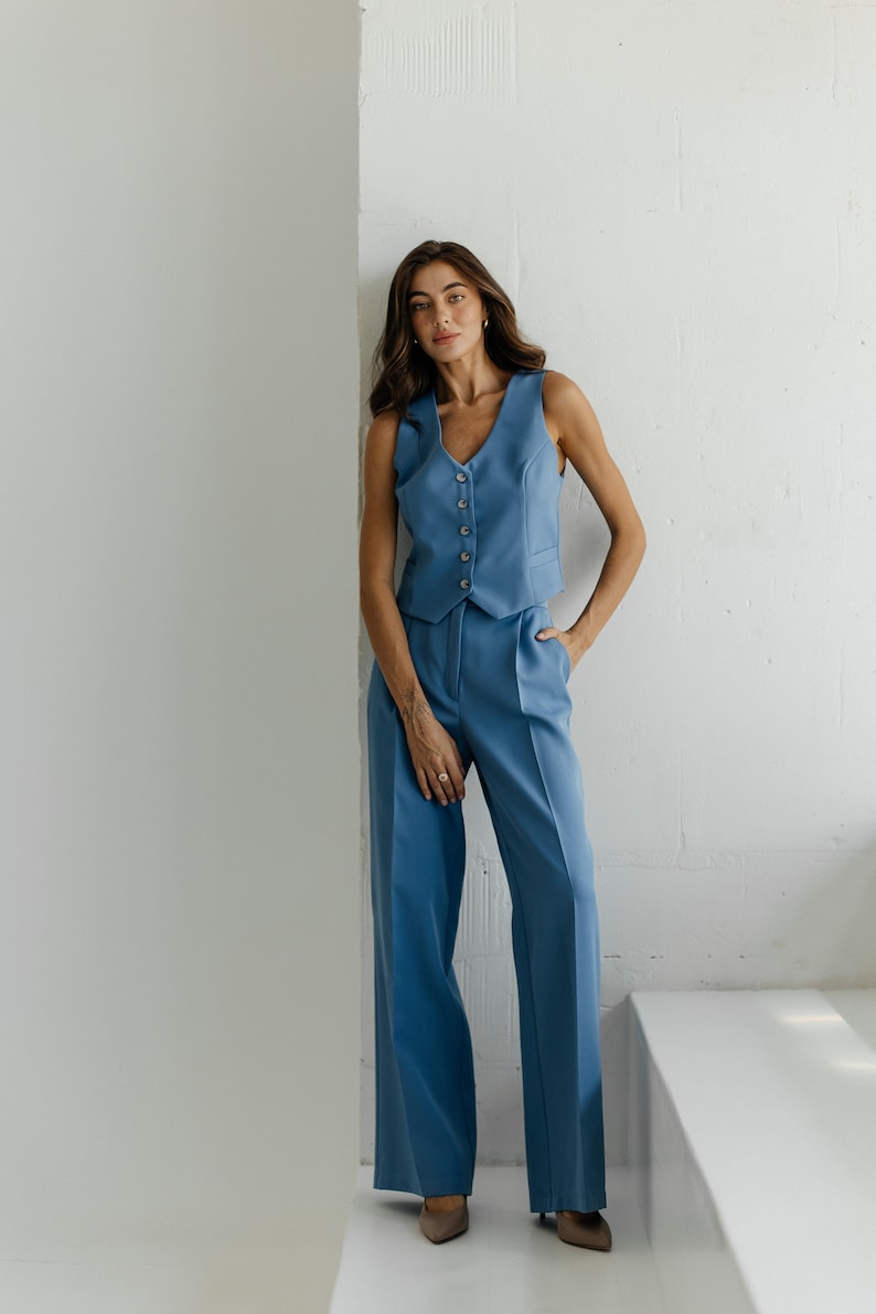 May include: A woman wearing a blue pantsuit with a vest and wide-leg pants. She is standing in front of a white wall.