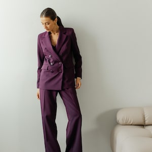 May include: A woman wearing a purple double-breasted blazer and matching pants. She is standing in front of a white wall with a light brown couch in the background.