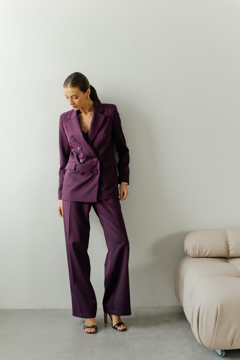 May include: A woman wearing a purple double-breasted blazer and matching pants. She is standing in front of a white wall with a light brown couch in the background.