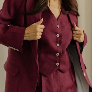 May include: A three-piece burgundy suit. The suit includes a blazer, a vest with button closures, and matching trousers. The blazer has a classic design with notched lapels and buttoned cuffs. The vest has a V-neck and a row of buttons down the front.