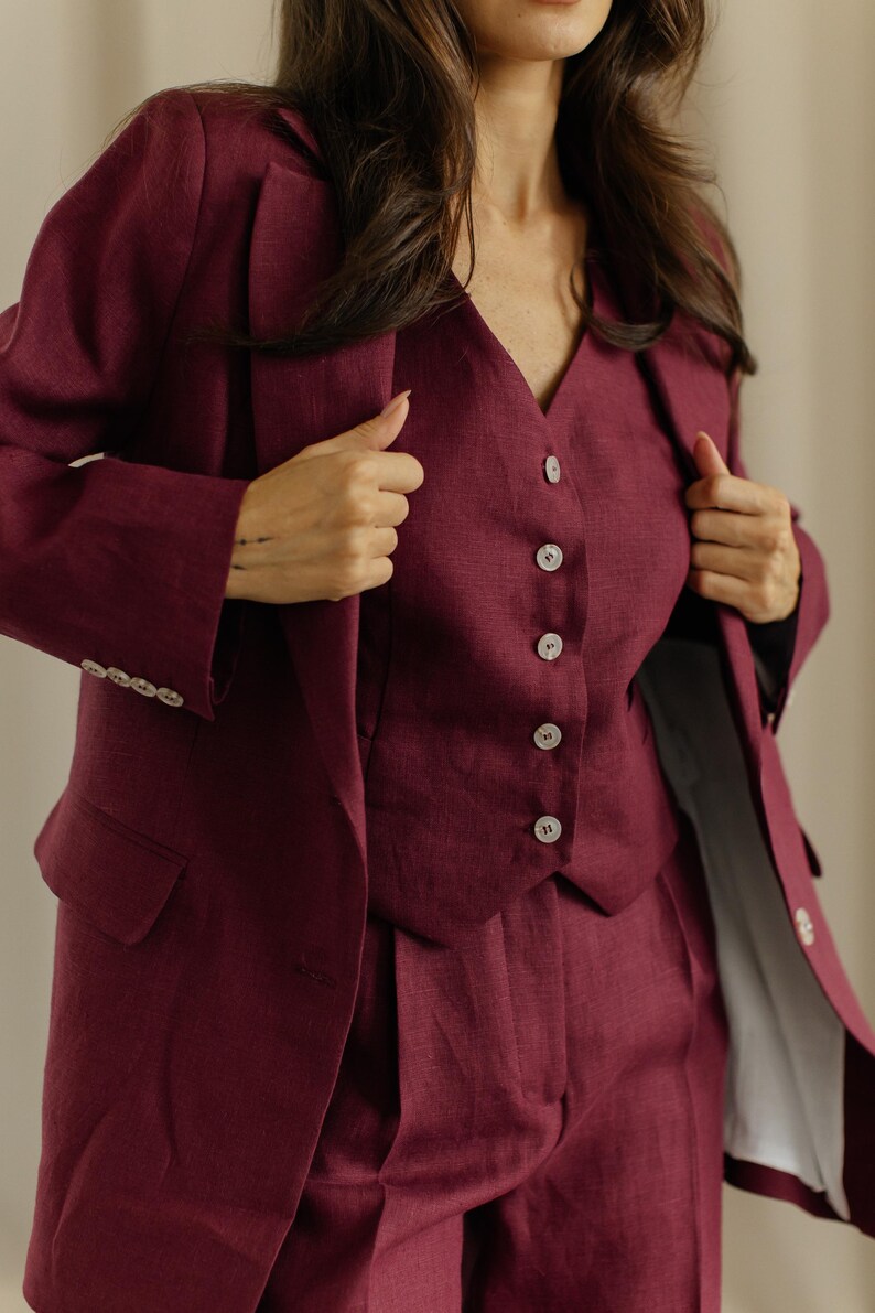 May include: A three-piece burgundy suit. The suit includes a blazer, a vest with button closures, and matching trousers. The blazer has a classic design with notched lapels and buttoned cuffs. The vest has a V-neck and a row of buttons down the front.